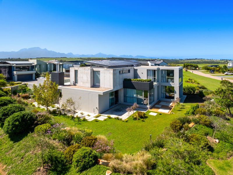 Aerial view of a modern architectural home featuring concrete finishes, solar panels, and extensive landscaped gardens in a scenic estate setting.