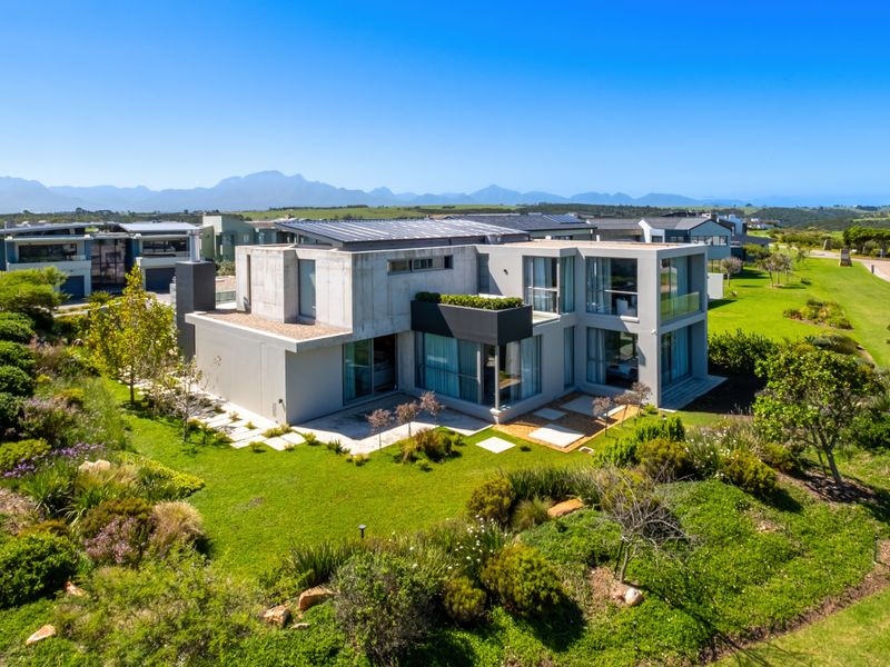 Aerial view of a modern architectural home featuring concrete finishes, solar panels, and extensive landscaped gardens in a scenic estate setting.
