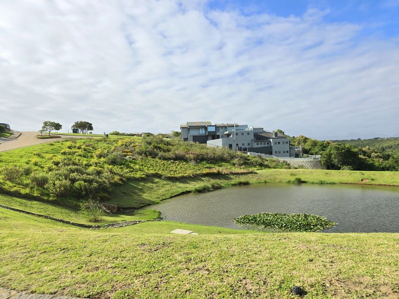 A scenic view of a residential plot featuring a natural pond, landscaped grass slopes, and surrounding estate architecture.