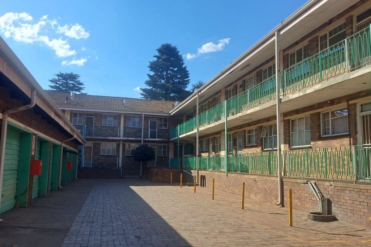 Exterior view of a multi-story apartment complex featuring brick facade, green metal balcony railings, and a paved courtyard area with garage units.