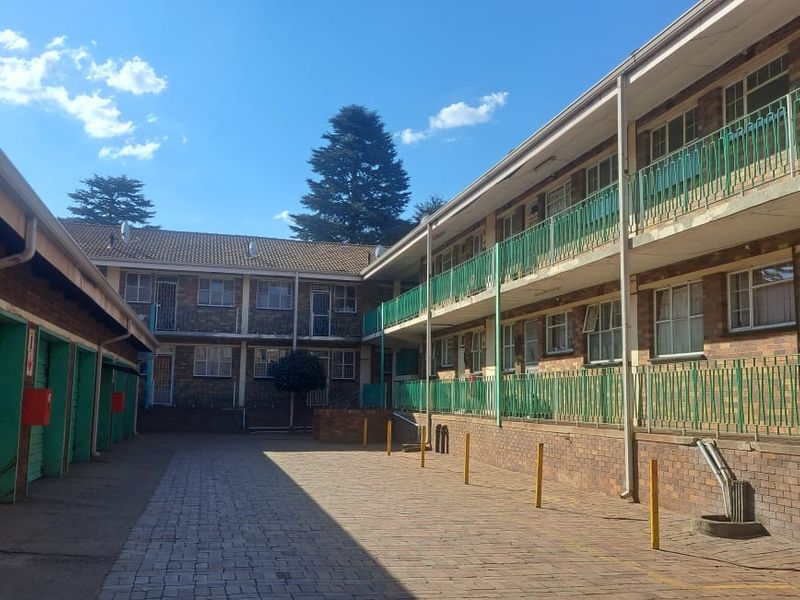 Exterior view of a multi-story apartment complex featuring brick facade, green metal balcony railings, and a paved courtyard area with garage units.