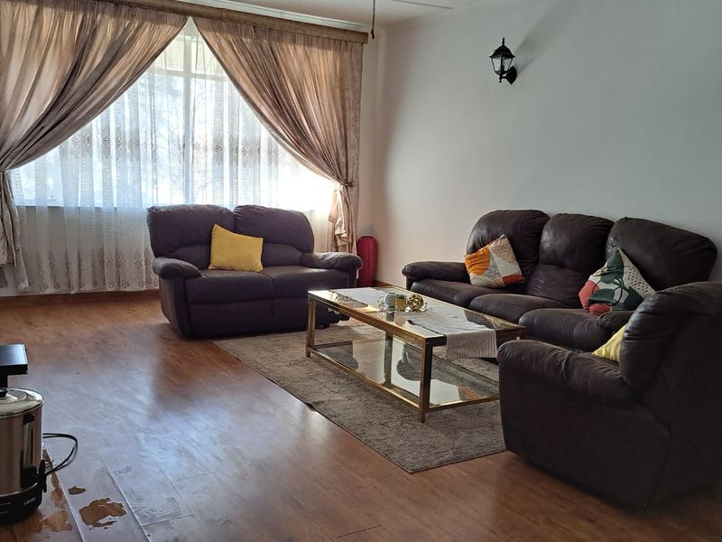 A spacious living room featuring wood-look flooring, large windows with curtains, and wall-mounted lighting fixtures.