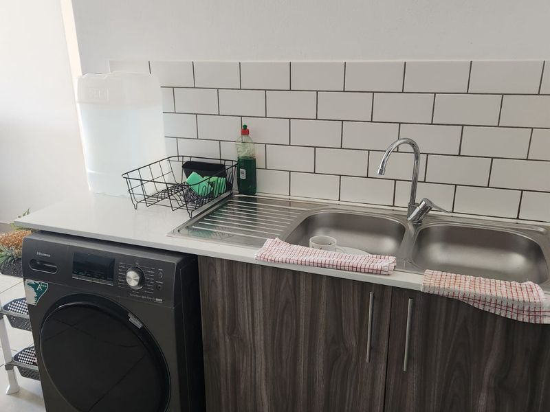 A functional laundry area featuring a stainless steel double sink, subway tile backsplash, and modern wood-grain cabinetry with a stone-look countertop.