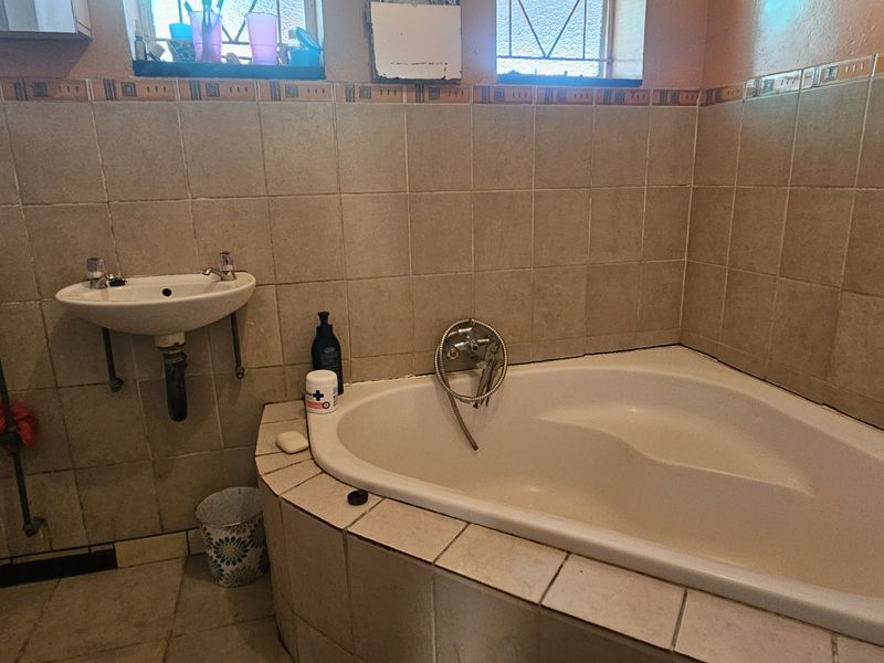 A dated bathroom featuring a corner bathtub, wall-mounted basin, and toilet, finished with neutral wall and floor tiles.