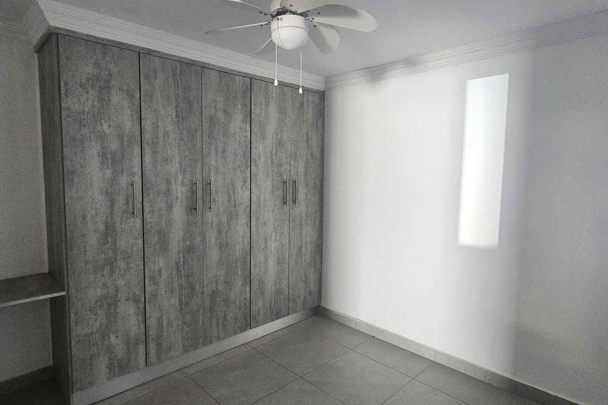 A clean, empty bedroom featuring modern grey wood-look built-in cupboards, tiled flooring, and a ceiling fan.