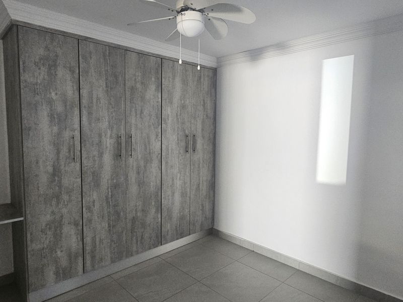 A clean, empty bedroom featuring modern grey wood-look built-in cupboards, tiled flooring, and a ceiling fan.