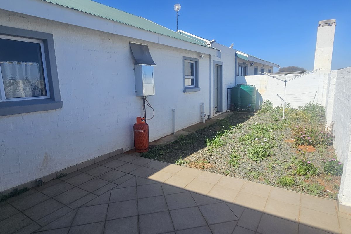 Rear exterior view featuring a tiled patio, boundary wall, water tank, and gas geyser installation with an adjacent garden area.