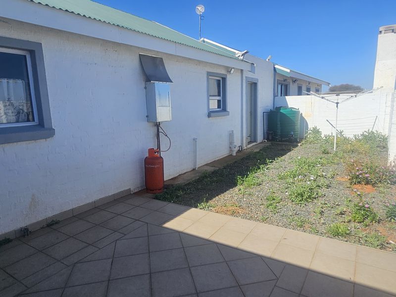Rear exterior view featuring a tiled patio, boundary wall, water tank, and gas geyser installation with an adjacent garden area.
