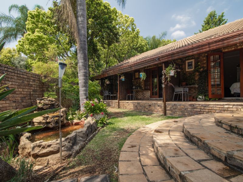 A charming rear garden area featuring a covered brick patio, stone-paved steps, and a decorative stone water feature set against a lush, mature garden.