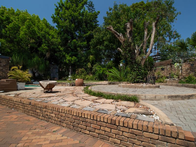 A spacious rear garden featuring a multi-level brick paved patio, retaining walls, and a swimming pool area surrounded by mature trees.