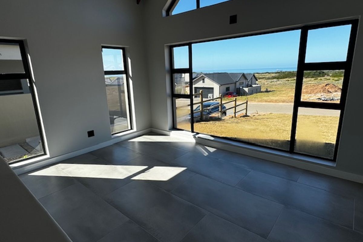 Spacious, light-filled living area featuring large format floor tiles, vaulted ceilings with exposed timber beams, and expansive windows offering scenic coastal views.