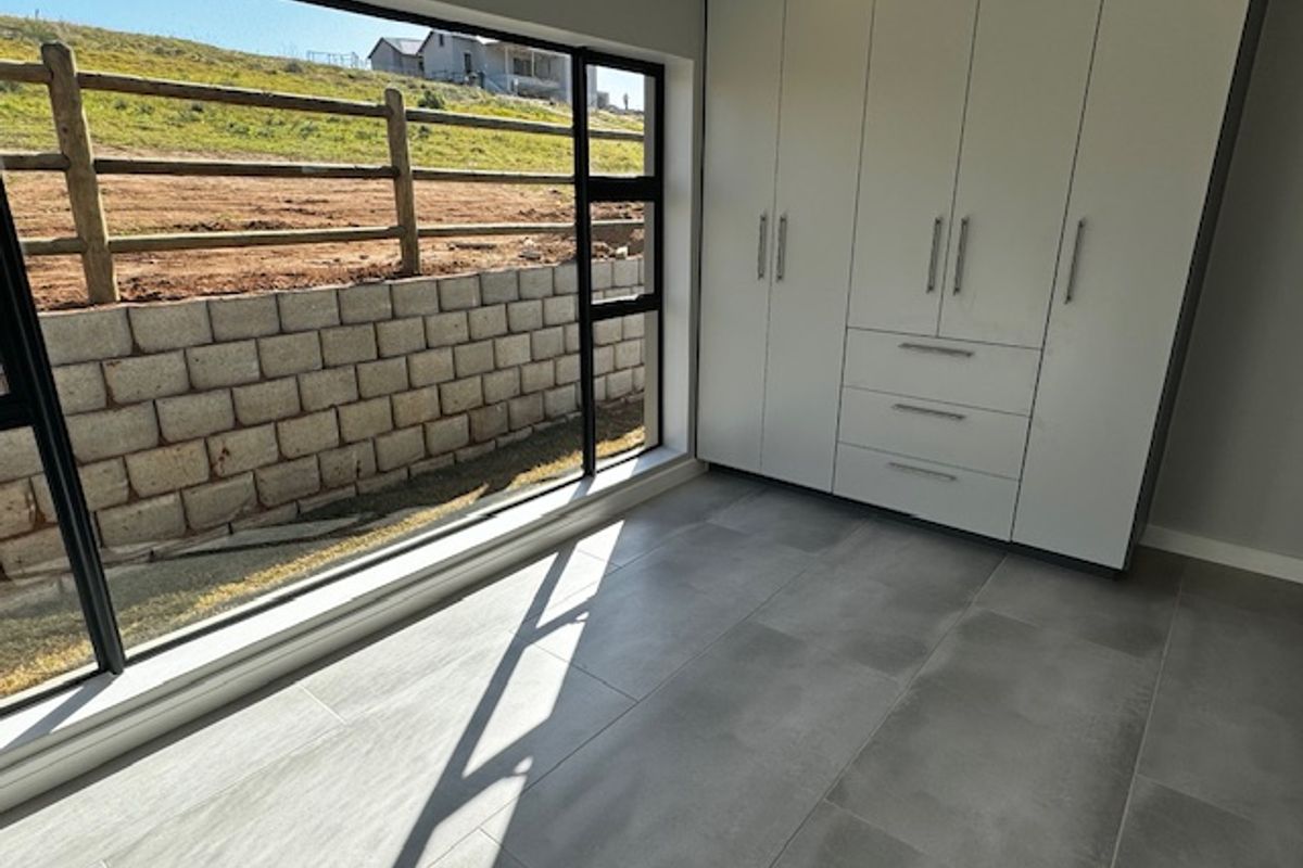 A modern, empty bedroom featuring large format floor tiles, built-in cupboards, and a large window with views of the exterior retaining wall.