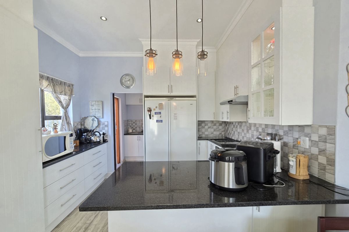 A well-maintained kitchen featuring white cabinetry, dark granite countertops, a tiled backsplash, and modern pendant lighting, with an adjacent scullery area.