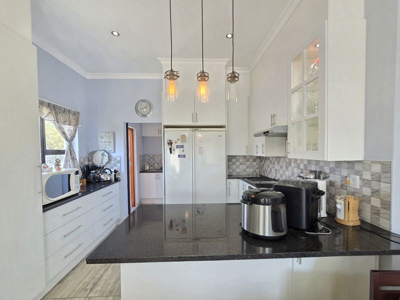 A well-maintained kitchen featuring white cabinetry, dark granite countertops, a tiled backsplash, and modern pendant lighting, with an adjacent scullery area.