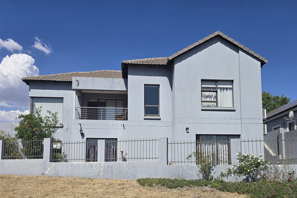 Rear view of a two-story residential house featuring a balcony, metal security gates, and a perimeter fence under a clear blue sky.