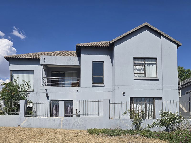 Rear view of a two-story residential house featuring a balcony, metal security gates, and a perimeter fence under a clear blue sky.