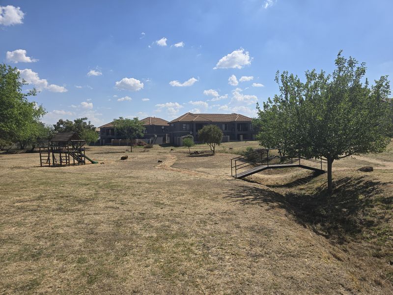 Expansive communal garden area featuring a pedestrian bridge, mature trees, and a children's play structure set against a residential complex background.