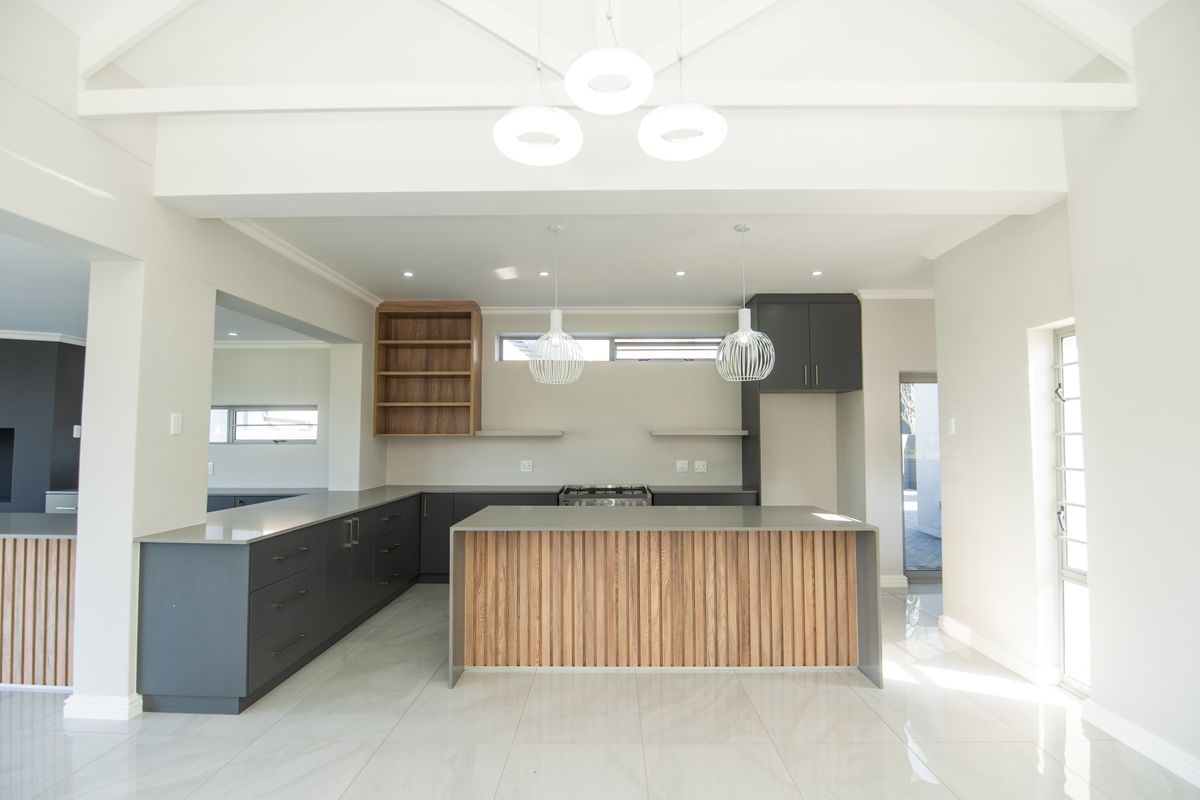 Modern, spacious kitchen featuring a central island, sleek dark cabinetry with wood accents, stone countertops, and high vaulted ceilings with contemporary pendant lighting.
