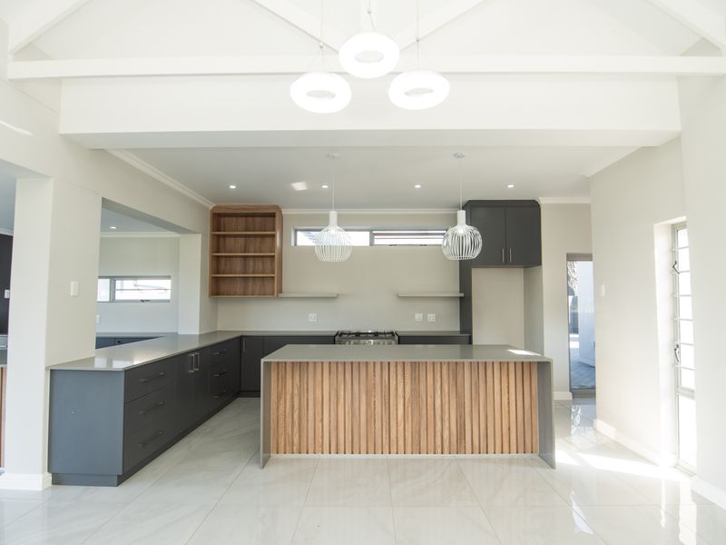 Modern, spacious kitchen featuring a central island, sleek dark cabinetry with wood accents, stone countertops, and high vaulted ceilings with contemporary pendant lighting.
