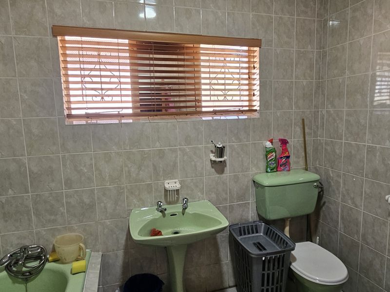 A dated bathroom featuring a green pedestal basin, matching toilet, and bathtub with full-height wall tiling and a window with venetian blinds.