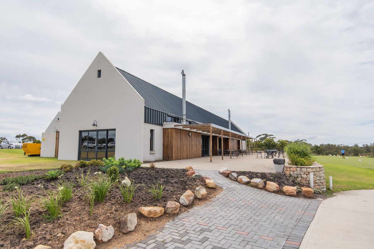 A modern farmhouse-style building featuring a metal roof, timber pergola, and a spacious paved patio area overlooking a landscaped garden.