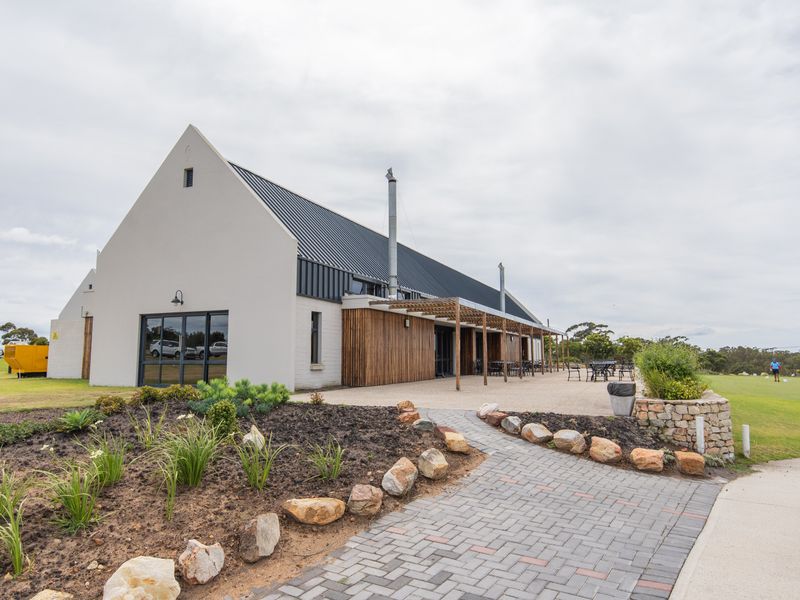 A modern farmhouse-style building featuring a metal roof, timber pergola, and a spacious paved patio area overlooking a landscaped garden.