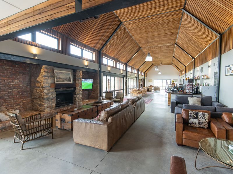 Spacious open-plan living area featuring a dramatic vaulted timber ceiling, exposed steel beams, polished concrete floors, and a large stone and brick fireplace.