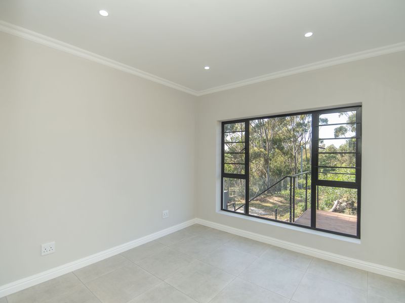 A bright, empty bedroom featuring neutral tiled flooring, recessed downlighting, and a large window with black aluminium frames overlooking the garden.