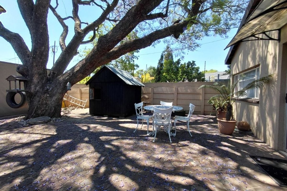 A rear garden area featuring a paved patio, a small storage shed, and a large mature tree providing shade over the outdoor space.