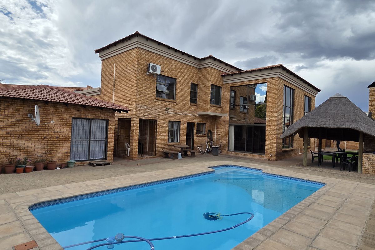 Rear exterior view featuring a swimming pool, paved patio area, and a traditional thatch roof lapa set against a double-story brick house.