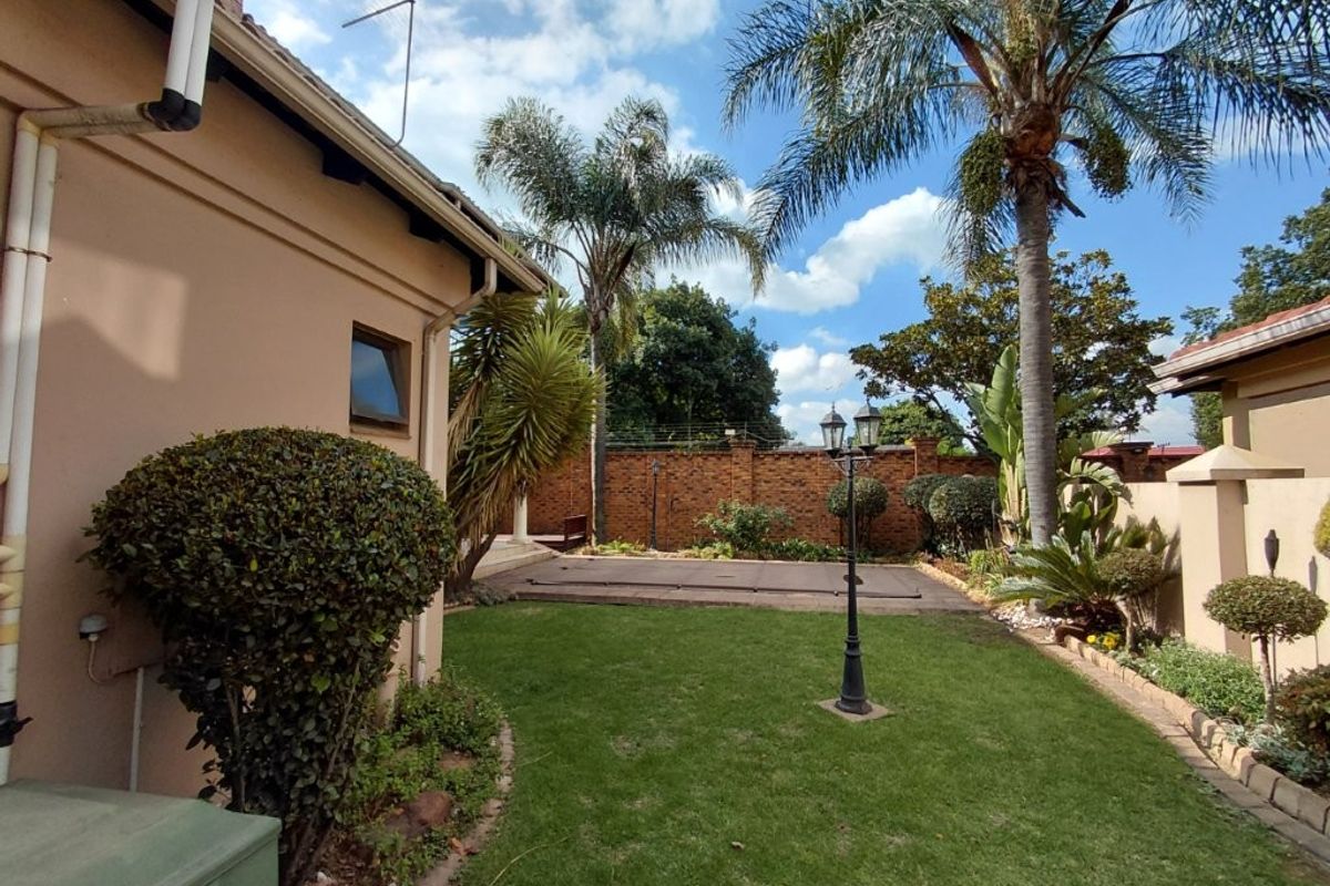 A well-maintained rear garden featuring a lawn, mature palm trees, brick boundary walls, and a covered swimming pool area.