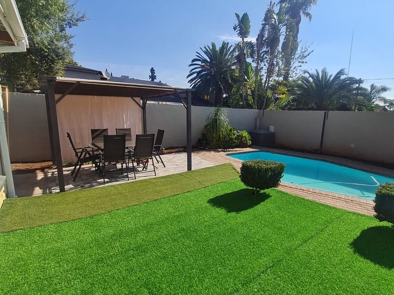 A well-maintained outdoor area featuring a swimming pool, a covered pergola patio, and a neat artificial lawn, enclosed by a boundary wall.