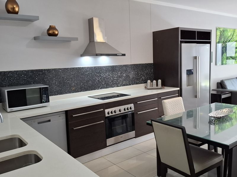 Modern open-plan kitchen featuring dark wood cabinetry, stone countertops, mosaic backsplash, and integrated stainless steel appliances, flowing into the dining and living area.