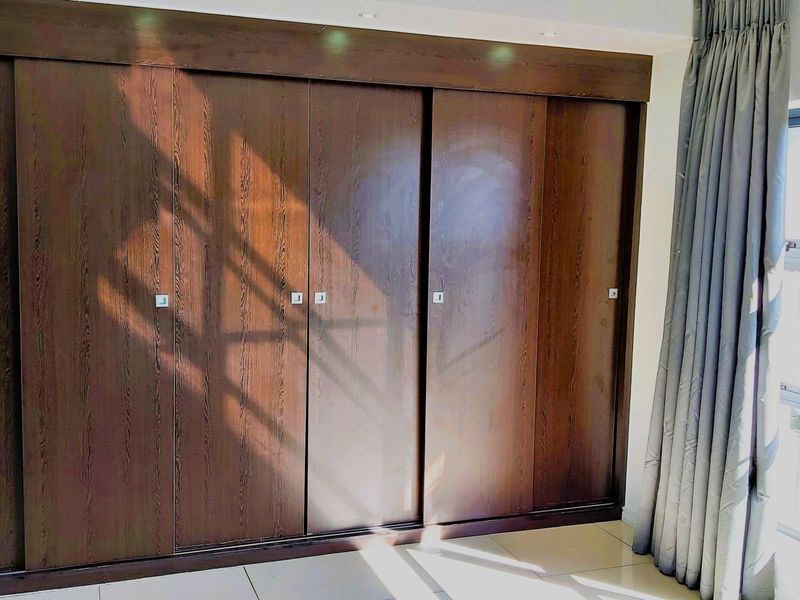 A bright bedroom featuring extensive dark wood built-in cupboards with sliding doors, neutral tiled flooring, and large windows with curtains.
