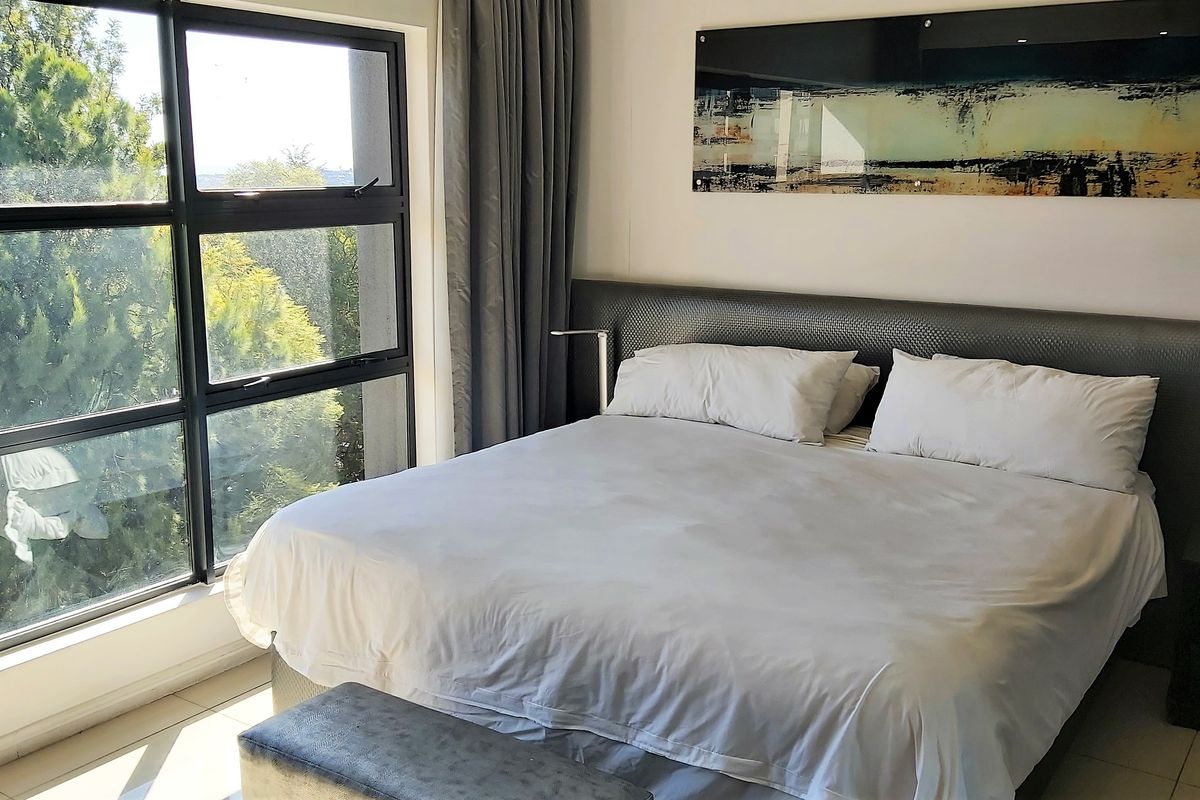 A well-lit bedroom featuring large aluminium-framed windows that provide natural light and views, complemented by tiled flooring and a modern built-in headboard.