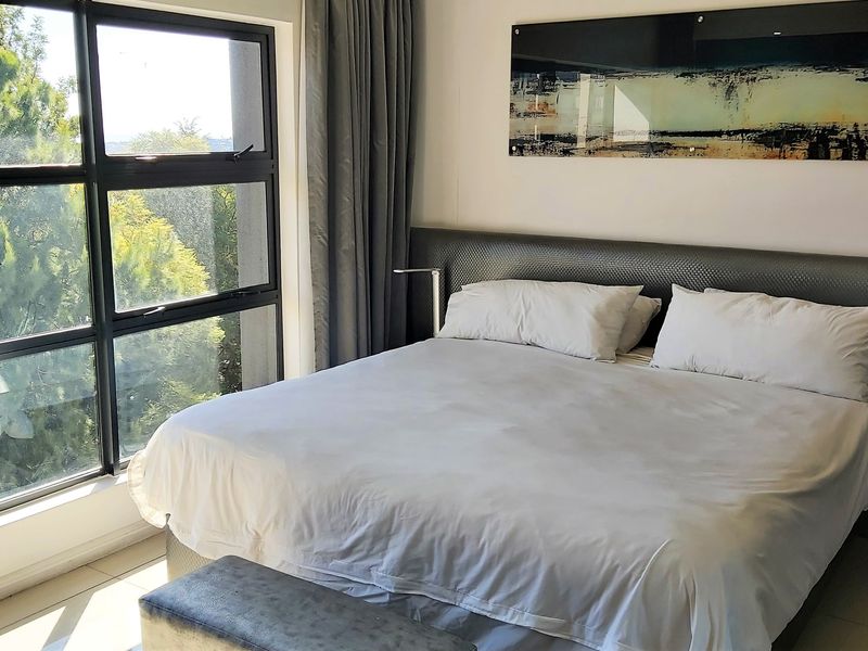 A well-lit bedroom featuring large aluminium-framed windows that provide natural light and views, complemented by tiled flooring and a modern built-in headboard.