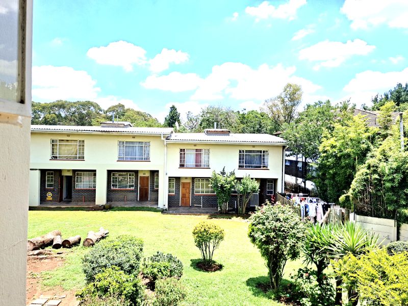 Rear view of a two-story apartment complex featuring a shared garden, brick and plaster facade, and metal-framed windows with security bars.