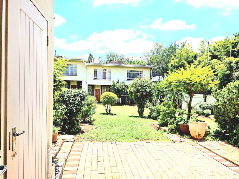 A view of a rear garden featuring a paved patio area and established landscaping, looking towards the back facade of the apartment building.