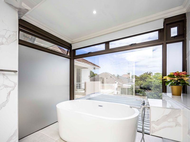 Modern bathroom featuring a sleek freestanding bathtub, marble-look wall tiles, and large windows offering natural light and neighborhood views.