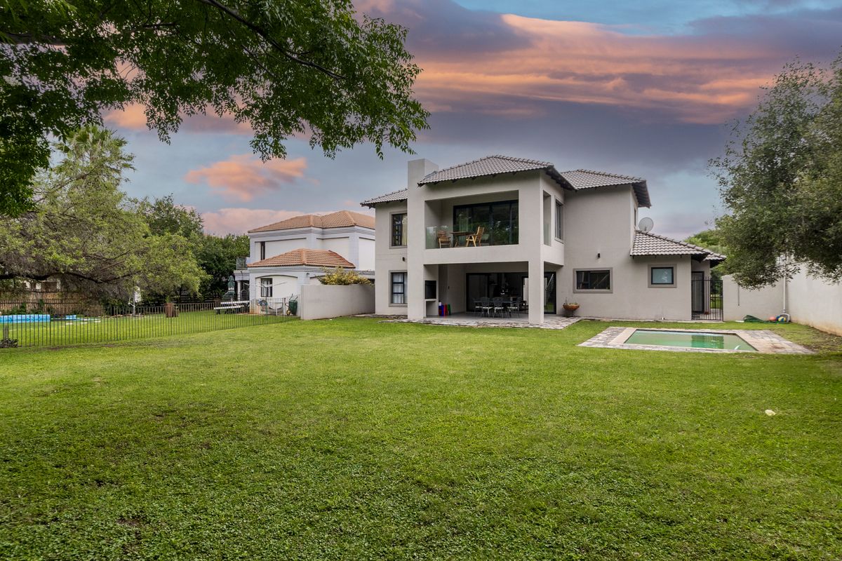Rear view of a double-story house featuring a spacious lawn, swimming pool, covered patio with built-in braai, and an upper-level balcony.
