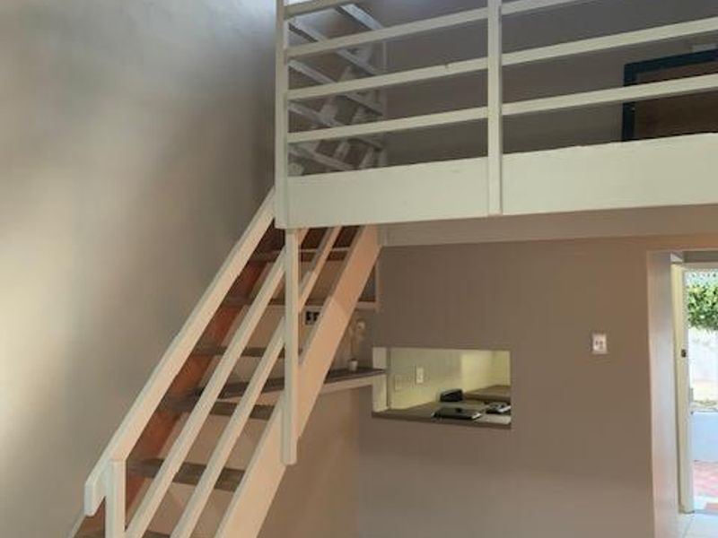 Open plan living area featuring a mezzanine level, exposed timber ceiling beams, and a staircase, with tiled flooring and a convenient pass-through window.