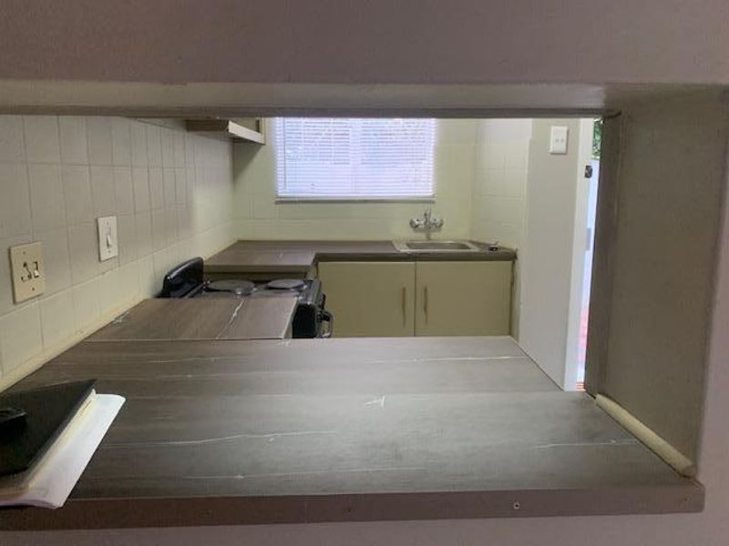 A functional kitchen featuring laminate countertops, tiled backsplash, built-in cabinetry, and a stainless steel sink with a view through a serving hatch.