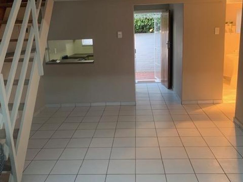 An open-plan living area featuring tiled flooring, a staircase leading to a mezzanine level, and a pass-through window connecting to the kitchen.