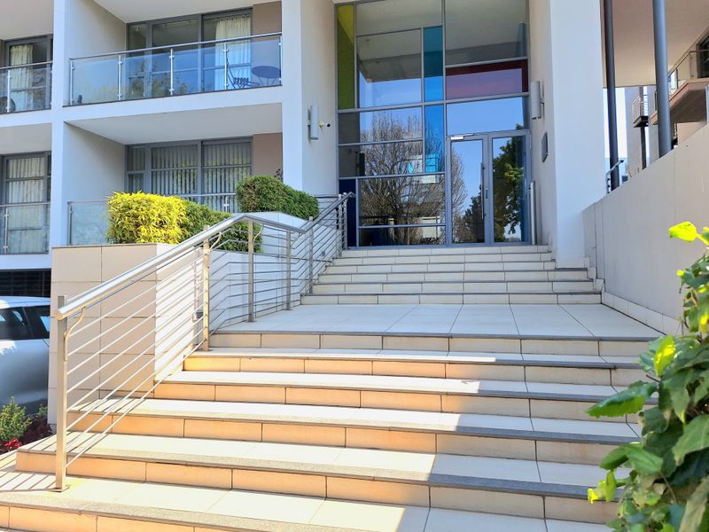 Modern apartment building entrance featuring a wide tiled staircase, stainless steel handrails, and a prominent glass facade with decorative panels.