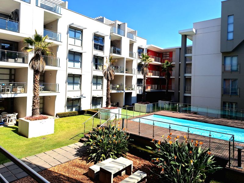 A well-maintained apartment complex courtyard featuring a swimming pool, timber decking, landscaped gardens, and modern glass-balustraded balconies.
