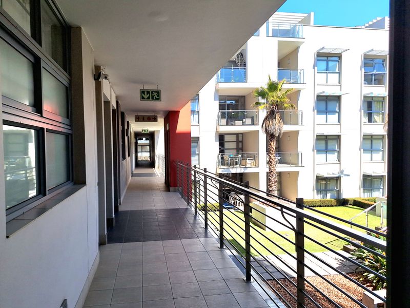 A well-maintained apartment complex hallway featuring tiled flooring, metal safety railings, and clear emergency signage, overlooking a landscaped courtyard area.