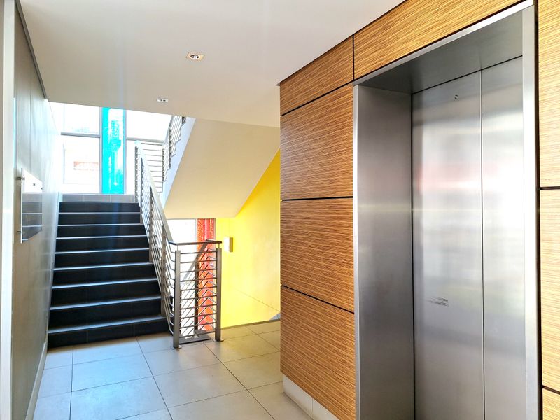 Modern apartment building hallway featuring a stainless steel elevator, tiled flooring, and a staircase with a metal balustrade.