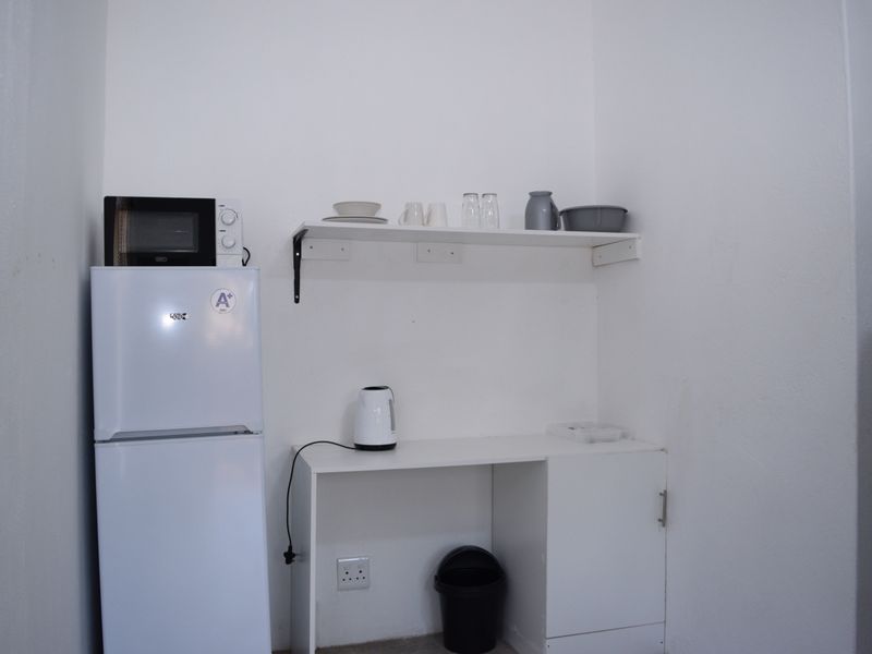 A compact kitchen area featuring basic white cabinetry, wall-mounted shelving, and dedicated space for a refrigerator.