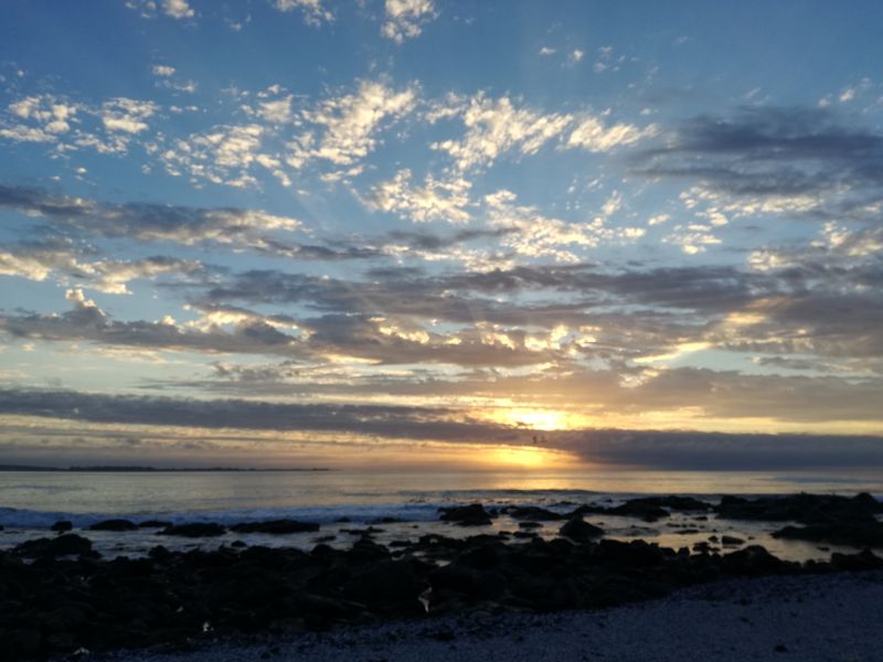 A scenic coastal view featuring a rocky shoreline at sunset with dramatic cloud formations over the ocean.
