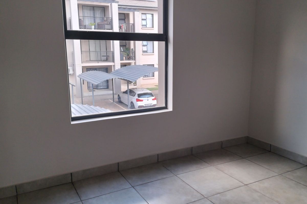 A bright, empty bedroom featuring neutral tiled flooring and a large window overlooking the apartment complex parking area.
